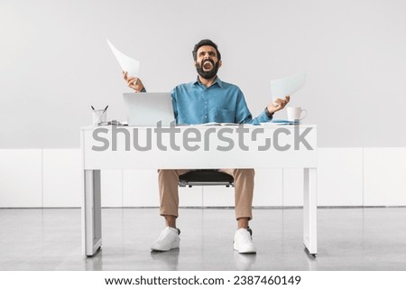 Overwhelmed indian businessman juggling multiple tasks, holding papers with distressed expression at clean, white office desk, full length shot Royalty-Free Stock Photo #2387460149