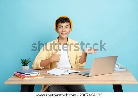 Portrait of handsome guy wearing stylish casual clothes and yellow hat, showing thumb up, looking at camera while sitting at desk with laptop. Education concept