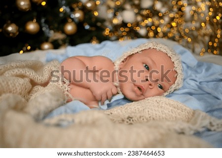 New Year Christmas holidays scene - cute newborn baby boy in funny hat lying on knitted beige blanket with Christmas festive tree gold toys and lights garland. Infant child items concept, Nursery