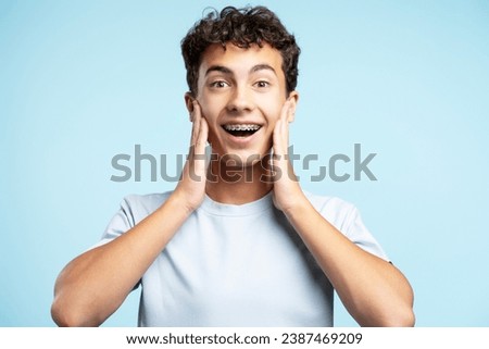 Surprised handsome teenage boy wearing dental braces touching face looking at camera isolated on blue background. Concept of positive lifestyle Royalty-Free Stock Photo #2387469209