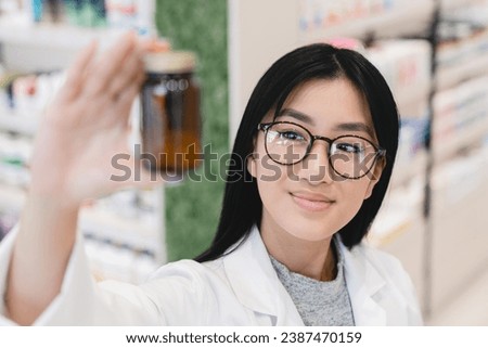 Asian young female druggist pharmacist in white medical coat looking at medicine drug jar, pills, remedies, painkillers, vitamins showing it to camera in pharmacy drugstore Royalty-Free Stock Photo #2387470159