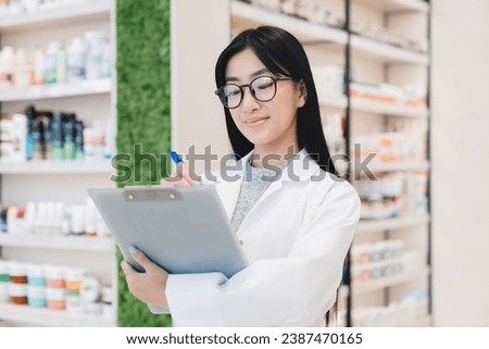 Asian female young pharmacist druggist in white medical coat writing on clipboard side effects, active substance, prescriptions standing at pharmacy drugstore Royalty-Free Stock Photo #2387470165