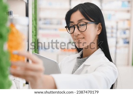 Closeup female asian pharmacist druggist in medical white coat looking for drugs at prescription stretching to pills, vitamins, painkillers, medicines at pharmacy shelves in drugstore Royalty-Free Stock Photo #2387470179