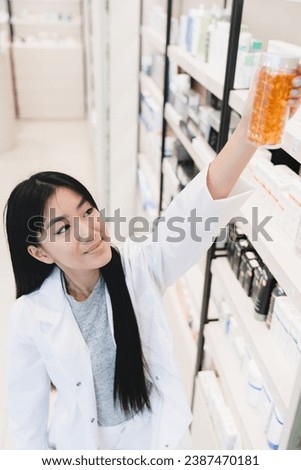 Female asian pharmacist druggist in medical white coat stretching to pills, vitamins, painkillers, medicines at pharmacy shelves in drugstore Royalty-Free Stock Photo #2387470181