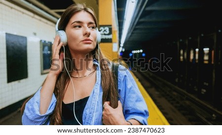 Young charming woman using mobile phone and listening music playlist with wireless headphones while waiting on tram station. City lifestyle concept. Travel in public transport. Female journey. Royalty-Free Stock Photo #2387470623