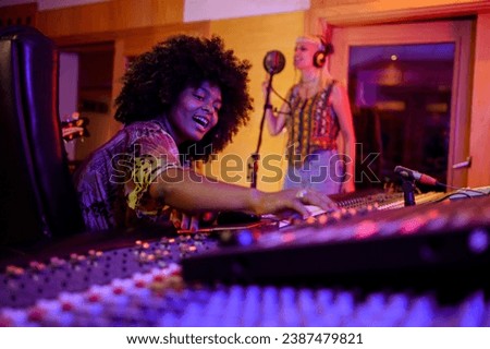 Portrait of a smiling multiracial female music producer operating her music console while female vocalist is being recorded. A happy sound technician is sitting in a recording studio and recording.