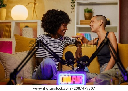Portrait of a two beautiful diverse female radio hosts recording podcast in a home broadcasting studio while sitting on a yellow sofa and talking to each other about feminine topics. Copy space.