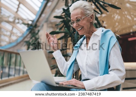 Elderly Caucasian woman in her 70s, smiling joyfully using laptop. She wear glasses, white shirt, , gesture with one hand, during video call. Indoor, daylight, concept of active seniors and technology