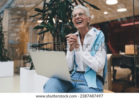 Exuberant elderly Caucasian woman in her 70s, with short white hair, laughs while using a laptop. She's possibly managing online investments or enjoying freelance work, seated in a co-working space Royalty-Free Stock Photo #2387487497