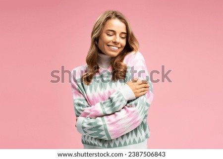 Attractive smiling woman wearing stylish comfortable winter sweater hugging herself  isolated on pink background, copy space. Cute fashion model posing for pictures. Shopping, sales, beauty concept
