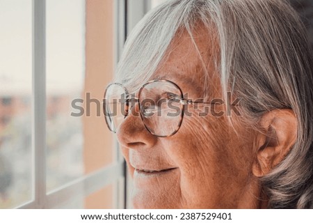 Dreamy smiling elderly 60s hoary woman looking in distance out of window, recollecting good memories or visualizing future at home, planning vacation or enjoying peaceful mindful day alone indoors. Royalty-Free Stock Photo #2387529401