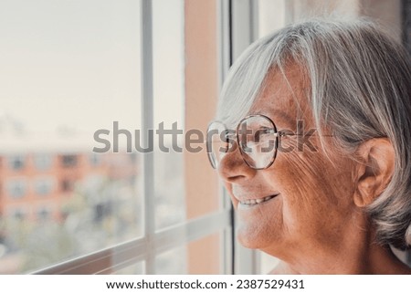 Dreamy smiling elderly 60s hoary woman looking in distance out of window, recollecting good memories or visualizing future at home, planning vacation or enjoying peaceful mindful day alone indoors. Royalty-Free Stock Photo #2387529431