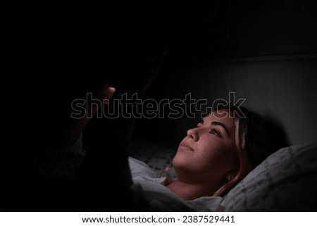 Gadget addiction. Calm smiling young woman or teenage girl lying at cozy bed holding cellphone in hands looking at screen chatting checking social network account before fall asleep at night