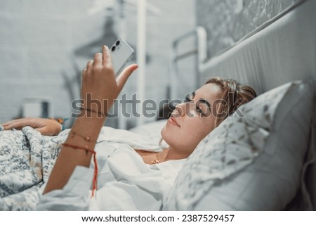 Gadget addiction. Calm smiling young woman or teenage girl lying at cozy bed holding cellphone in hands looking at screen chatting checking social network account before fall asleep or after waking up
