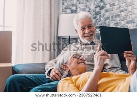 Happy retired senior family couple relaxing on sofa after moving activities, sharing tablet computer, looking at screen, laughing, making video call, using online app, Internet service