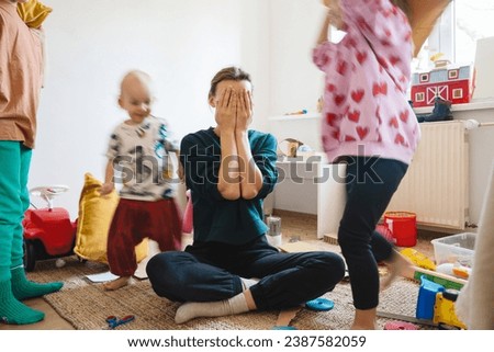 Stressed out mother sitting on floor in middle of toys while children naughty running around her at room. Woman alone burnout with kids. Family home with chaos, mess. Motion blur for speed, real life. Royalty-Free Stock Photo #2387582059