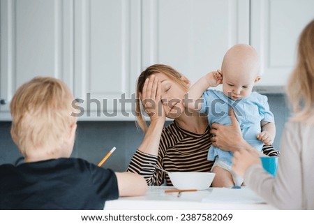 Young exhausted woman with three children at home. Tired sleepy mother with baby on her lap drinking coffee while her older children do homework or drawing in kitchen at home. Motherhood burnout. Royalty-Free Stock Photo #2387582069