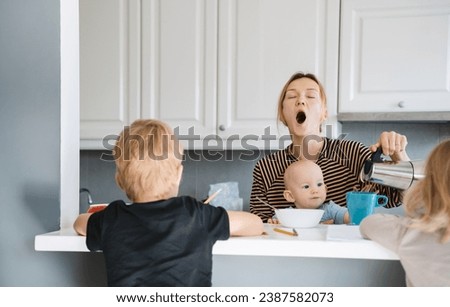 Young exhausted woman with three children at home. Tired sleepy mother with baby on her lap drinking coffee while her older children do homework or drawing in kitchen at home. Motherhood burnout.