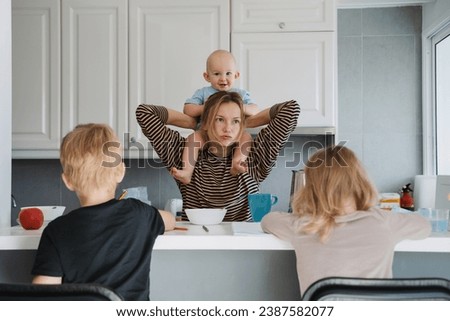 Young exhausted woman with three children at home. Tired sleepy mother taking care of baby while her older children do homework or drawing in kitchen at home. Motherhood burnout.