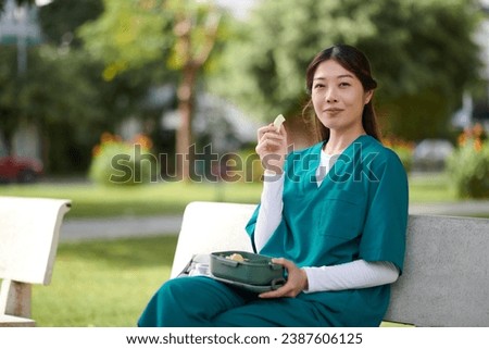 Portrait of cheerful medical nurse eating fresh fruits when resting on bench in park during her break Royalty-Free Stock Photo #2387606125