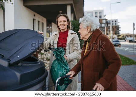 Woman helping elderly neighbor throw away trash into garbage can, waste container in front of their apartment complex. Royalty-Free Stock Photo #2387615633