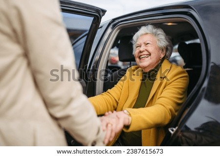 Senior lady getting out of the car, caregiver helping her, holding her hands. Elderly woman has problem with standing up from the car back seat. Royalty-Free Stock Photo #2387615673