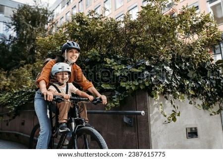 Mother carring her son on a child bike carrier, seat, both wearing helmets. Mom commuting with a young child through the city on a bicycle. Royalty-Free Stock Photo #2387615775
