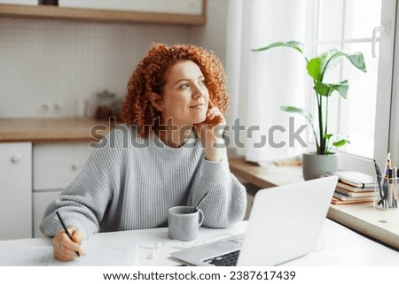 Pensive dreamy female with red curly hair sitting at kitchen table looking aside writing in her diary. Pretty student girl thinking about her dream job doing homework in front of laptop Royalty-Free Stock Photo #2387617439