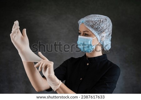 Caucasian young woman doctor or nurse in black uniform and protective mask putting protective glove isolatedon dark. Healthcare concept