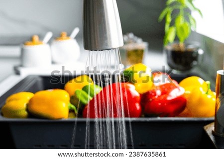 Yellow and red pepper are washed in a special tray for vegetables. The stream of water pours on the pepper in the sink. The interior of a modern kitchen.