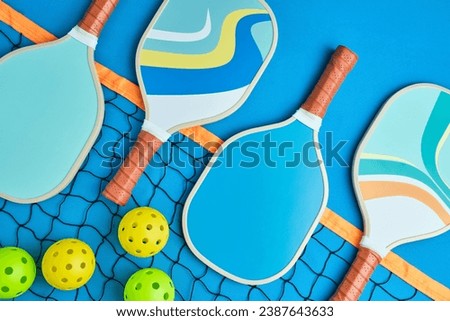 four pickleball paddles in blue tones on top of a net on a blue background with three balls next to it Royalty-Free Stock Photo #2387643633