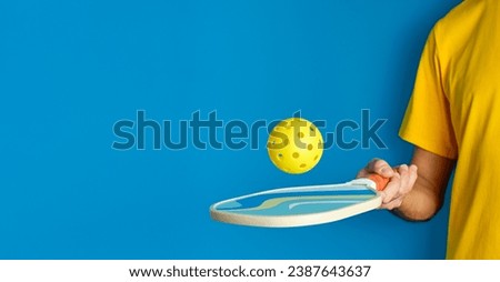 hand with paddle and yellow pickleball ball on blue background Royalty-Free Stock Photo #2387643637