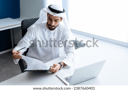 Handsome man with dish dasha working in his business office of Dubai. Portraits of a successful businessman in traditional emirates white dress. Concept about middle eastern cultures. Royalty-Free Stock Photo #2387660401