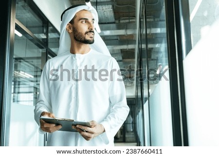 handsome man with dish dasha working in his business office of Dubai. Portraits of a successful businessman in traditional emirates white dress. Concept about middle eastern cultures. Royalty-Free Stock Photo #2387660411