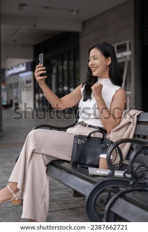Side view image of a beautiful and confident millennial Asian woman or businesswoman is talking on a video call on her phone while resting on a bench in the city. People and technology concepts