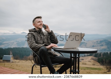 Workplace in country on backyard house in morning. Man talking on phone outdoors. Male work on a laptop sitting table with great view mountains. Concept remote work or freelance lifestyle. Internet 5G Royalty-Free Stock Photo #2387668839