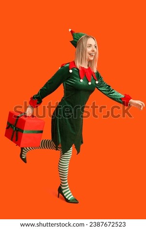 Young woman in elf costume holding Christmas gift box on orange background