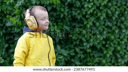 A positive little boy of 6 years old in a yellow jacket and yellow retro earphones listens to music. The boy stands against the background of a wall of plants. Banner with space for text