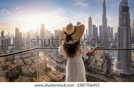 A woman in a white dress and hat looks at the panoramic sunset view of the downtown district skyline of Dubai, UAE Royalty-Free Stock Photo #2387706453