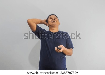 An astonished young Asian man wearing a navy t-shirt tries to switch channels, finds a less pleasantvchannel, becomes unpleasant when the viewing on TV is not suitable, isolated on a white background