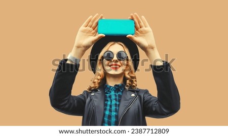 Portrait of stylish happy laughing woman stretching her hands for taking selfie with mobile phone wearing black round hat, leather jacket and sunglasses on gray background