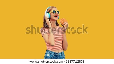 Portrait of happy laughing young woman listening to music in headphones holding burger fast food on yellow studio background