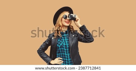 Portrait of beautiful young woman photographer with film camera taking picture wearing black round hat, leather jacket on brown studio background