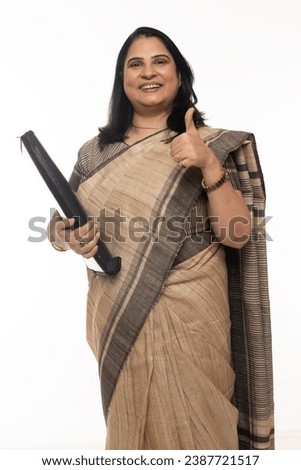Portrait of Indian lady teacher showing thumbs up and wearing in saree  while holding folder. Confident traditional Indian woman against white background