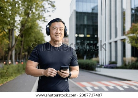 Smiling young Asian man standing on city street, running wearing headphones and holding phone, looking away.