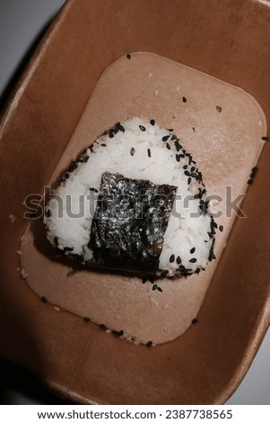 Onigiri in a cardboard food delivery box. Dark background, strong flash, closeup of onigiri details, onigiri covered in sesame seeds. Funny and creative food picture. Asian food culture. Takeout food.