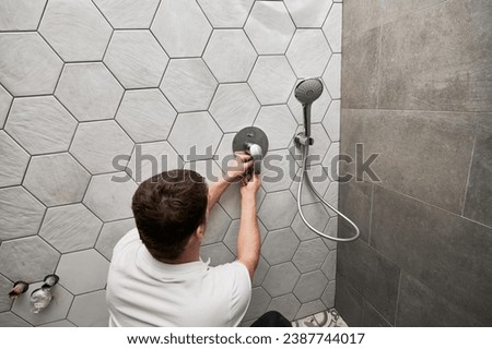 Back view of male plumber installing wall mounted shower system in apartment under renovation. Man repairing metal shower faucet handle while working on bathroom renovation. Plumbing works concept. Royalty-Free Stock Photo #2387744017