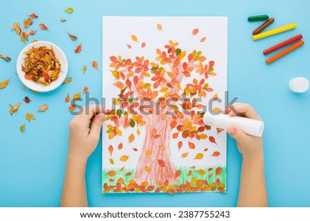 Little child hands gluing and creating tree shape from colorful leaves on white paper. Light blue table background. Pastel color. Making autumn decorations. Point of view shot. Closeup.