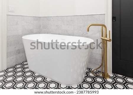 A freestanding tub with a gold faucet, black and white pattern tile flooring, marble subway tile backsplash, and a black sliding door. Royalty-Free Stock Photo #2387785993