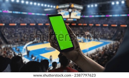 Basketball Championship: Person's Hand Holding a Smartphone with a Green Screen Display. Sports Fans in an Arena Cheering for a Team to Win. Template to Use for Social Media, Scores, Results, Betting